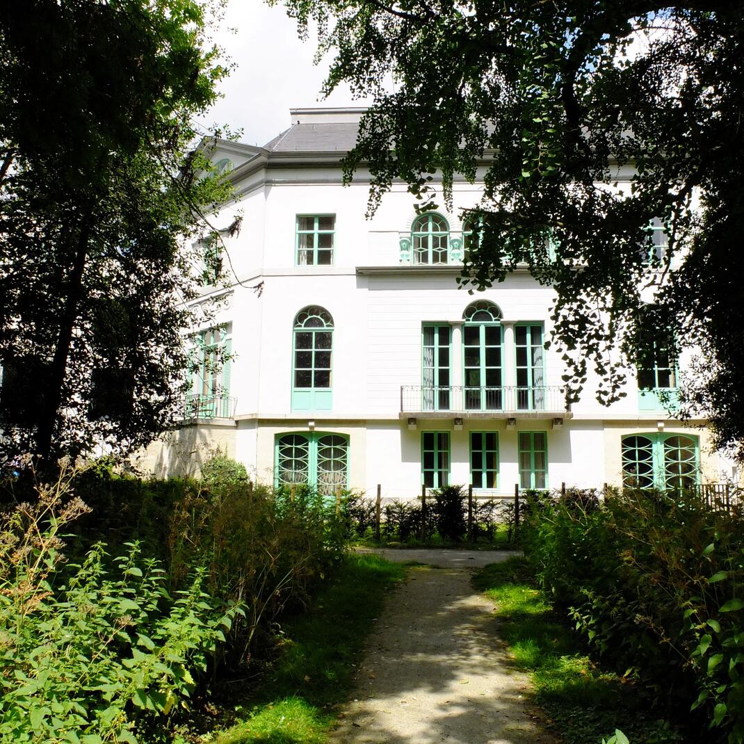 Een blik op de achtergevel van de witte villa, door de bomen heen.