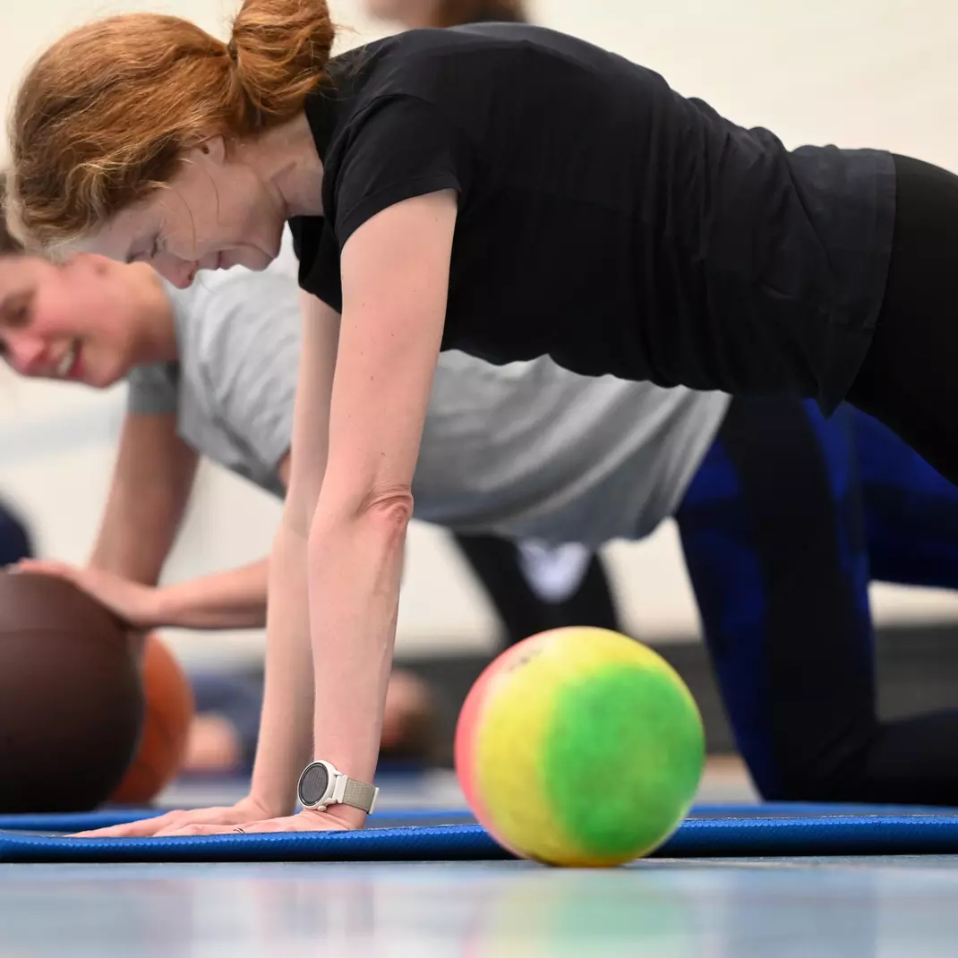 Twee dames die fitoefeningen met behulp van een bal op een matje doen in een sporthal