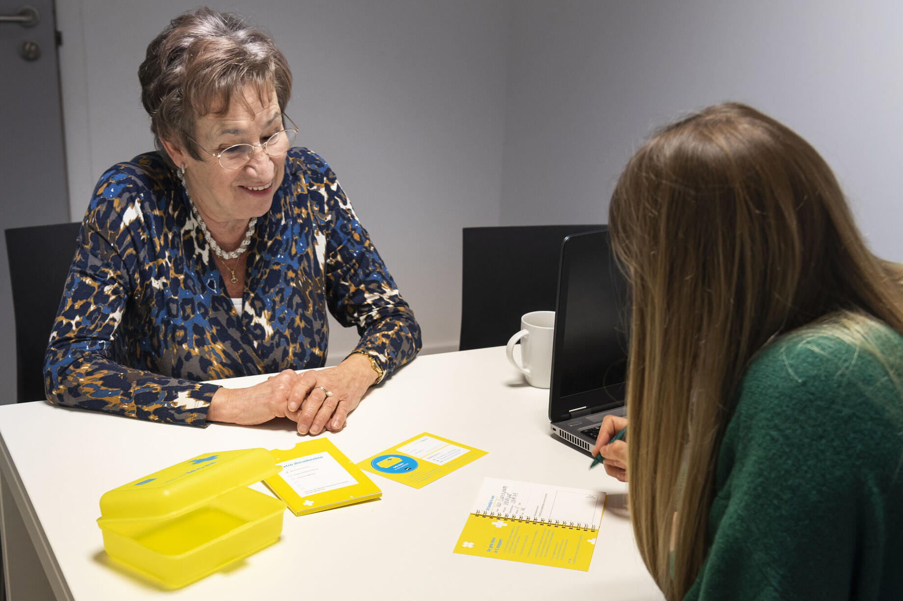 een mevrouw aan een tafel met hulpverlener, die de gele doos invult